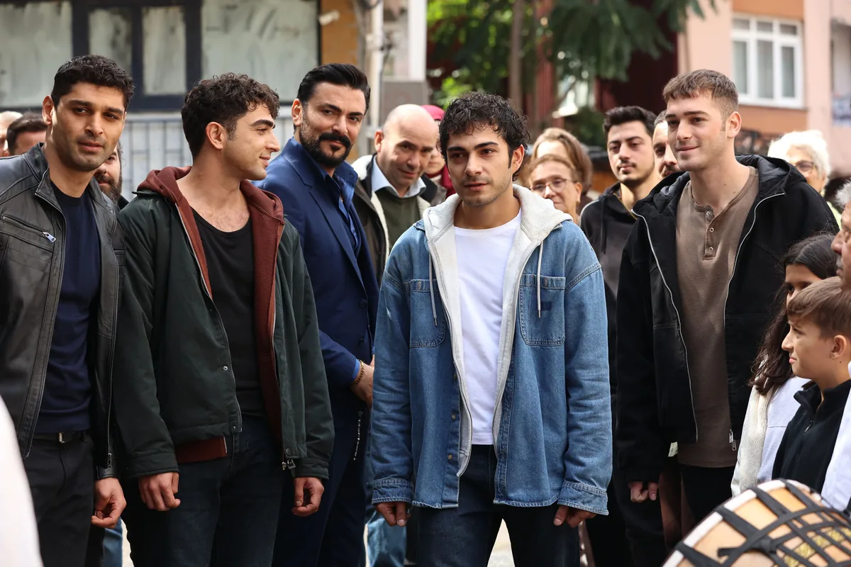 Altı Üstü İstanbul dizisinden, mahalle sokağında yan yana duran genç erkekler ve arkalarındaki kalabalık